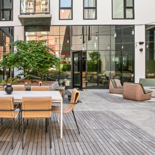 Outdoor courtyard at The Brady featuring dining tables, woven lounge seating, string lights, landscaping, and modern apartment façades.
