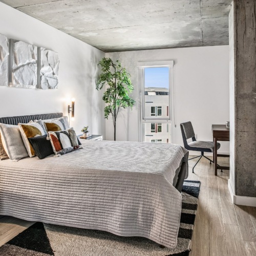 Bright bedroom at The Brady with an exposed concrete ceiling, wood-style flooring, large window, upholstered bed, and modern furnishings.